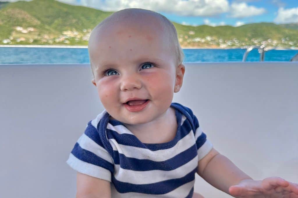 A baby boy in a blue and white striped t-shirt is looking at the camera and smiling. This is a close up.  You can see in the back ground the sea and an island. His family are sailing with him when he is a baby. 