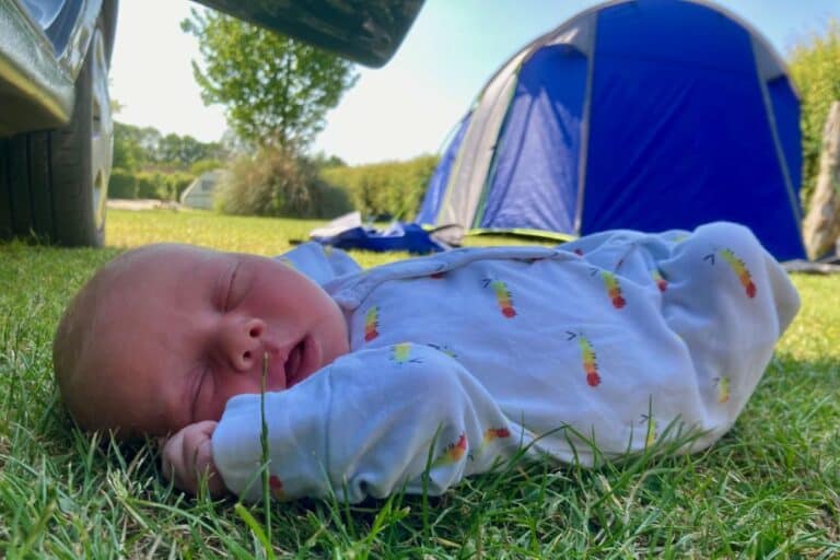 A newborn baby is lying on the grass in his white and patterned onsie with his eyes shut. In the background is a tent. This is the families first camping trip with their baby.