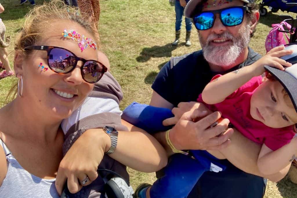 A woman is at a festival and wearing sunglasses and gems on her face in silver and has her baby over her shoulder facing away from the camera. Her boyfriend is next to her wearing sunglasses and baseball cap and holding their toddler son who has on a pink t-shirt and baseball cap. They are looking at the camera as they are having a photo taken as they love family friendly festivals in the UK where they are.