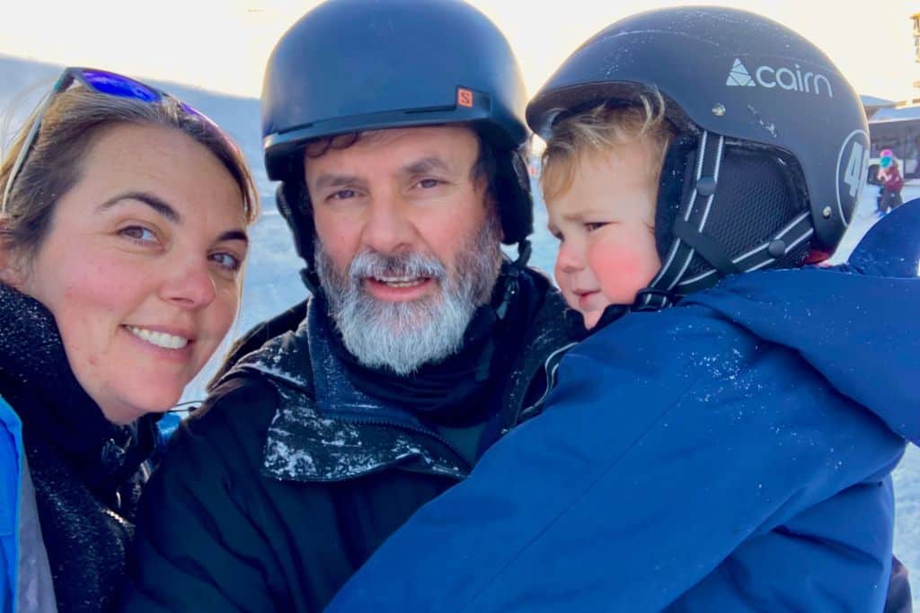 A woman is smiling at the camera with sunglasses on her head. Next to her is her boyfriend who has on a ski helmet, and ski jacket. He is holding their toddler who that are skiing with.