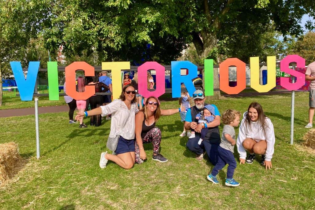 A family is crouching down in from of a 3D sign saying "Victorious" as they are at Victorious Festival in the UK. It is one of the best family friendly festivals.
