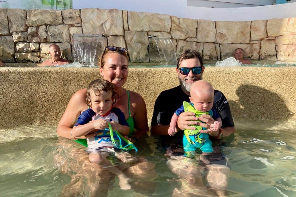 A mother and father are sat in a  shallow and small swimming pool. On her lap she has her toddler son and on his lap he has his baby son. They are all looking at the camera smiling.