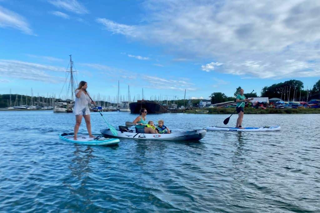 There is a woman in a kayak with two boys sat in the front of the kayak. They are on the River Hamble. Next to her are two other women and each of them is on a stand up paddle board either side of her in her kayak. They are kayaking with the kids to the pub. In the background is marina and lots of yachts can be seen.