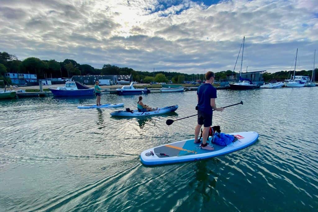 You can see the backs of a group of people on the water either kayaking or stand up paddle boarding. To the left is a man on a paddle board with a dog on the board. In the middle is a woman kayaking with two kids, and on the left is another woman stand up paddling. In the background is a marina. They're in the River Hamble.