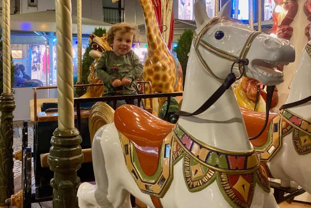 There is a fairground carousel and there is a little boy riding in a horse carriage. in the front of his is a white horse that is pulling his cart. He's smiling at the camera a like he's having a great time. 
