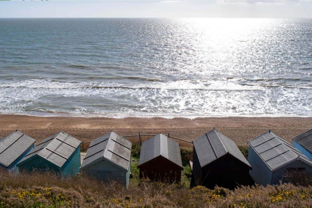 In the foreground is the back of a row of beach huts and as you look down on them from a cliff edge.  In front of the beach huts is a shingle and sand beach and then the sea.  The sun is reflecting on the sea.