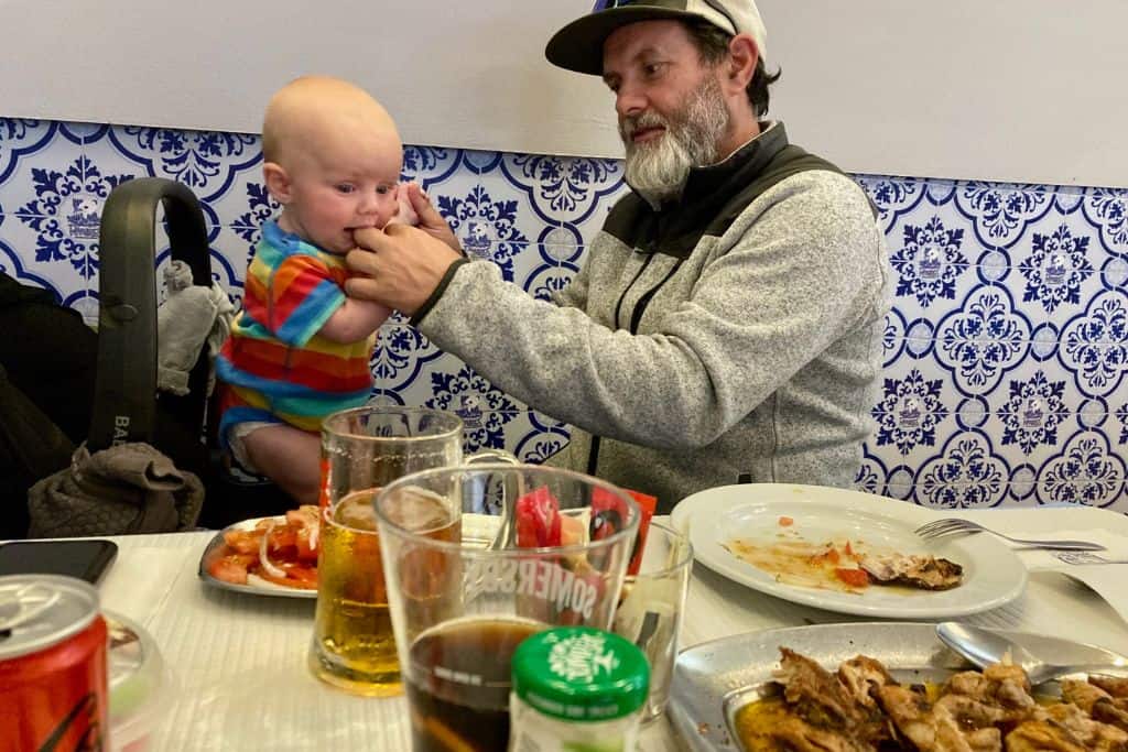 A man is side on wearing a baseball cap and a fleece.  Opposite is a baby in a multicoloured striped onesie. The man is holding the babies hands as he stands up in his car seat. In the front of the image is a table and on it are various dished and some drinks. Behind them are blue and white tiles on the wall. They are eating dinner in the Algarve.