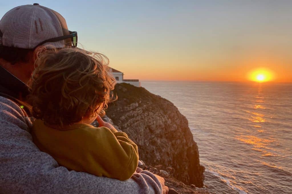 There is a man with his back to the camera and on his lap is his toddler age son. They are sat on a cliff above the sea and looking over to the right as the sun is looking to set into the sea. 