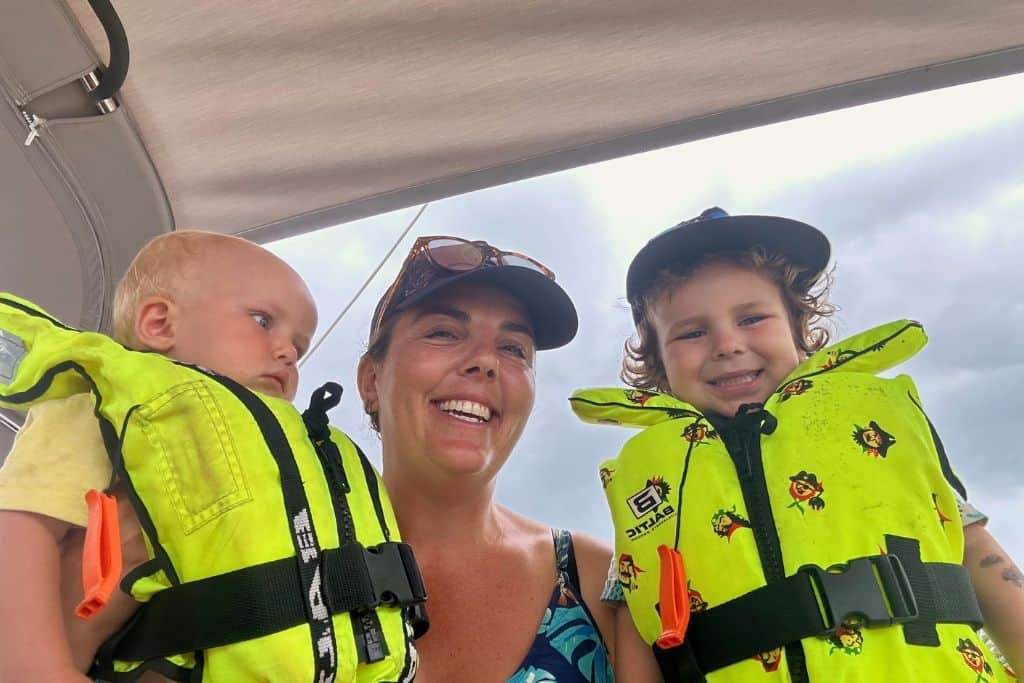 In the middle of the image is a woman looking at the camera. On her left is her three year old son. On her right is her baby son. The boys both have on yellow lifejackets. She is sailing with her baby for the first time. 