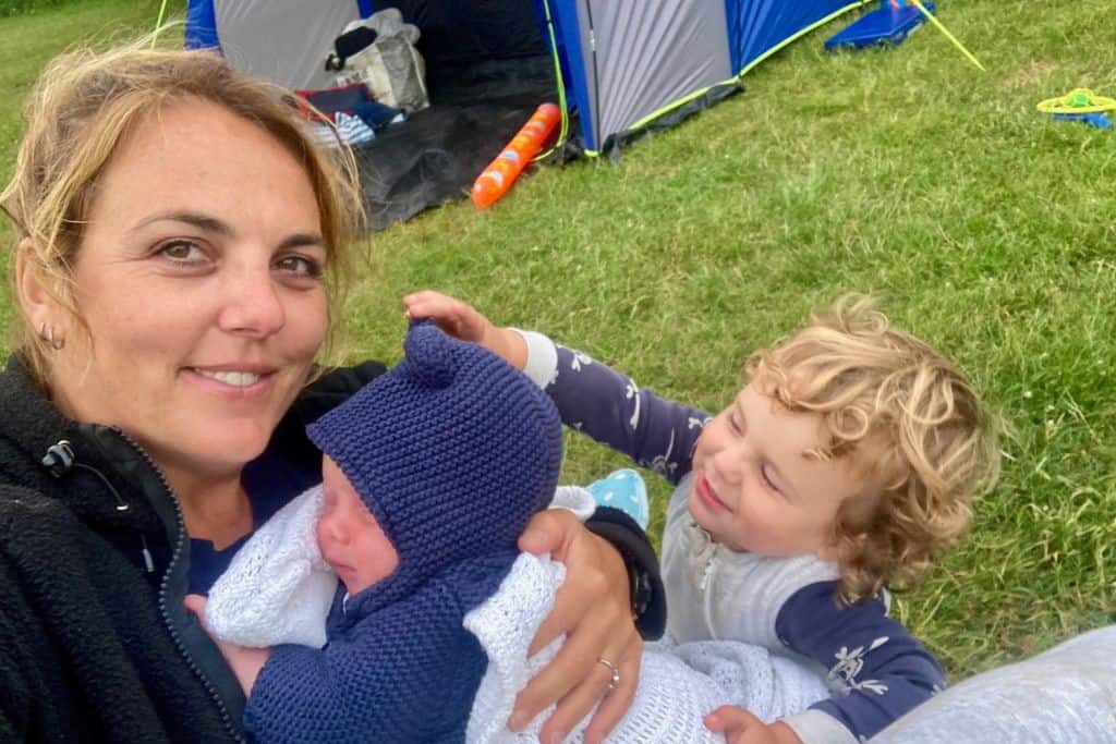 A woman is sat in a camping chair in a field looking at the camera and has her baby in a blue knit hoodie and in a blanket on her lap. Stroking the babies head is his brother. In the background is a tent and she's camping with her kids.