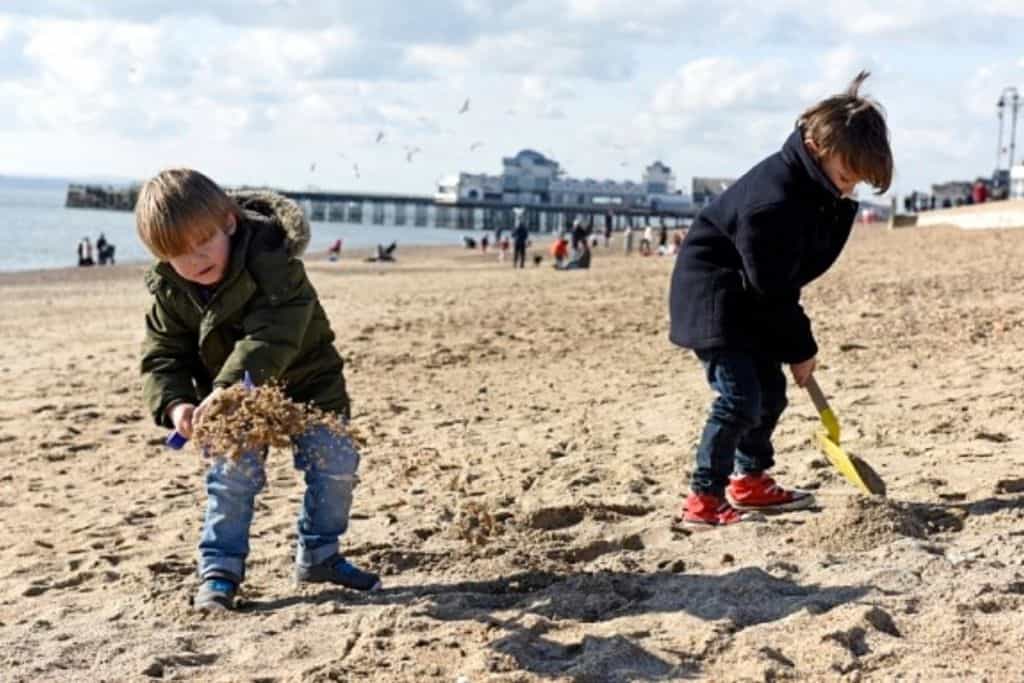 There is a sand beach and on it are who young boys in front of the camera in winter coats playing with spade and the dry seaweed.  In the backgound behind them is the pier. This is Southsea Beach which is one of the sandy beaches in Hampshire.