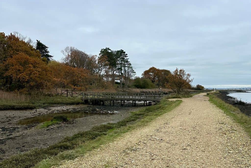 There is a foot path in the centre of the image and to the left is a small marsh area with a bridge going across it. In the distance are some trees. This path is part of a walk from Hamble Le Rice to Warsash.