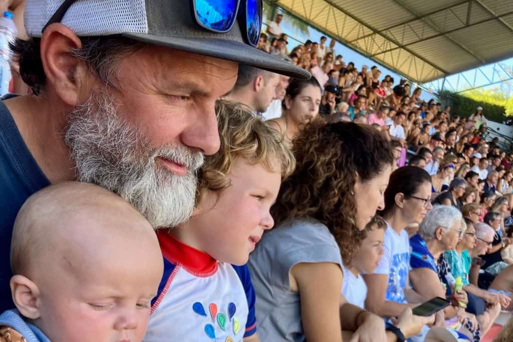 A man with a baseball cap and sunglasses resting on top of it is looking to the right. He is watching a show at Zoomarine. Next to him on his left is his toddler son who is also watching the show. On his right at the front of the image is his baby son who is looking away from the show. 