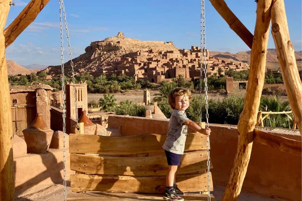 A little boy is stood on a wooden swing and looking at the camera. Behind him in the background is Ait Benhaddou a UNESCO site in Morocco.