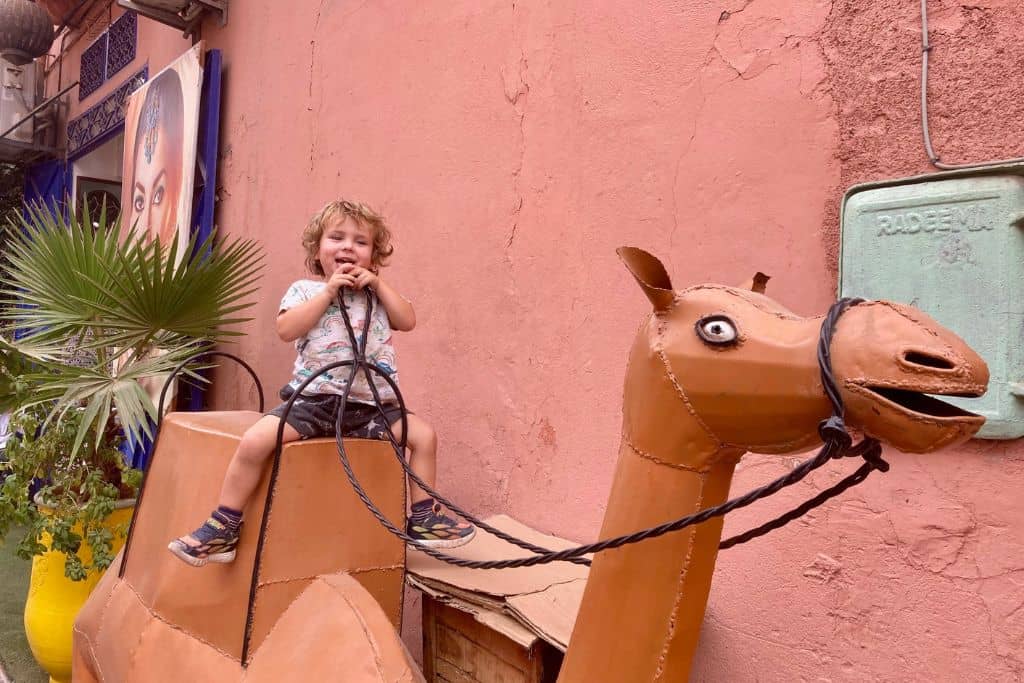 A three year old boy in shorts and t-shirt is sat on a kneeling statue of a camel in the souk in Marrakech.