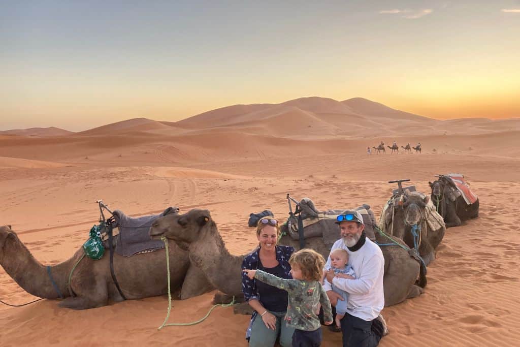 The photo is taken in the Erg Chebbi sand dunes. In the front of the image are three camels lying down on the sand and in front of them is a family. There is a mum wearing a blue shirt kneeling down next to and looking at her son. Her son is pointing off to the left and talking about something. Next to the woman also kneeling is her boyfriend who is in a white shirt and holding their baby son. 