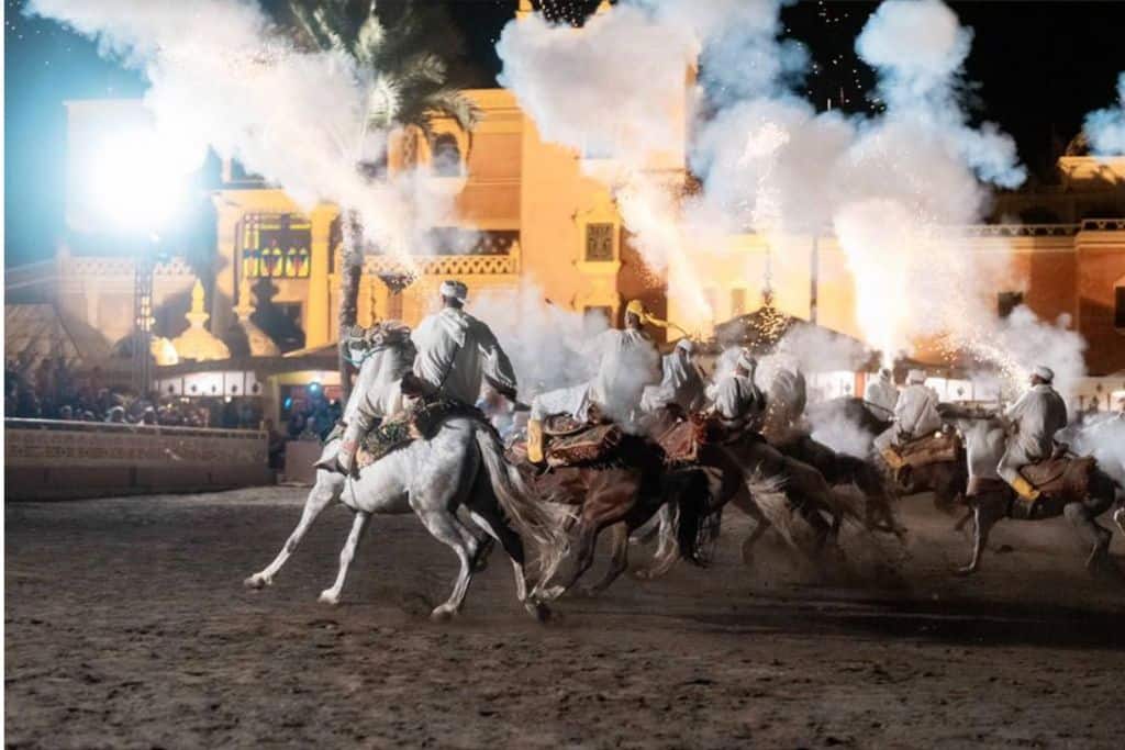 This it the Chez Ali Fantasia Show in Marrakech. There are some men riding white horses letting of smoke flares as the horses perform.