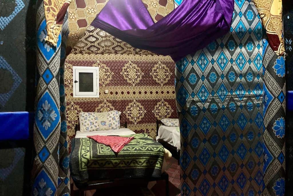 This is the inside of a luxury desert camp in the Erg Chebbi sand dunes in the Sahara in Morocco. The image is taken looking through to the bedroom.  There are lots of brightly coloured fabrics on the walls and on the bed.