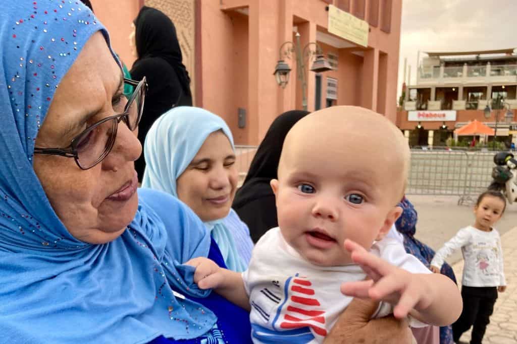 A baby in a white t-shirt with a sailboat on it is smiling at the camera. He is being held by a Moroccan lady a blue hijab and wearing glasses. She is smiling at the baby. Behind them are more local women in blue as well. This is in Ouazazate in Morocco where the baby's parents are staying with their kids.