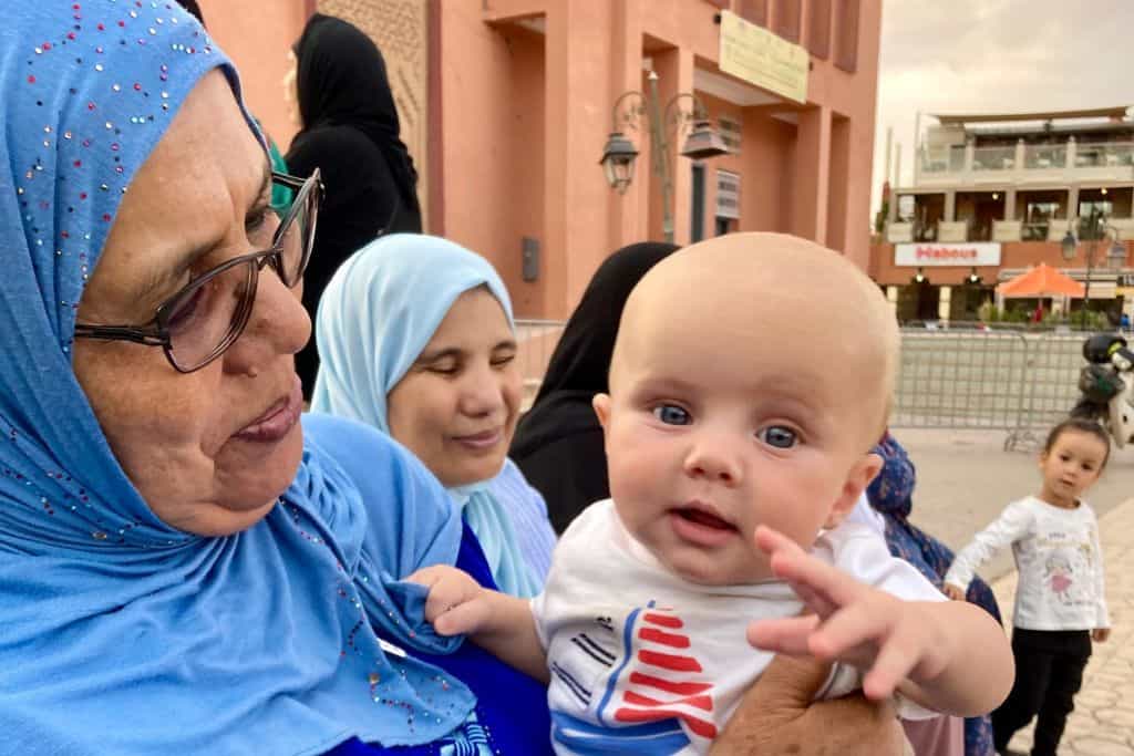 A baby is looking at the camera as the photo is being taken. He is being held by a local woman wearing a blue hijab which is what the women choose to wear in Marrakech. She has on glasses and is smiling at him.