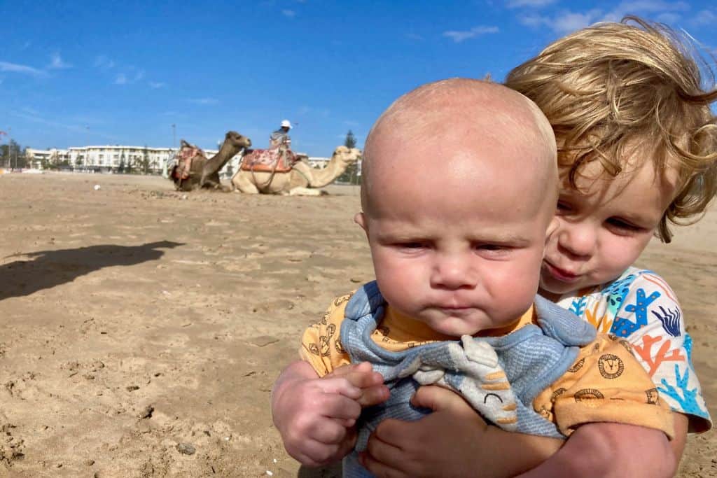 A baby is sat looking at the camera and his 3 year old brother is cuddling him from behind and smiling at the camera.  They are to the right of the image and to the left behind them is the beach with some camels sat on it.