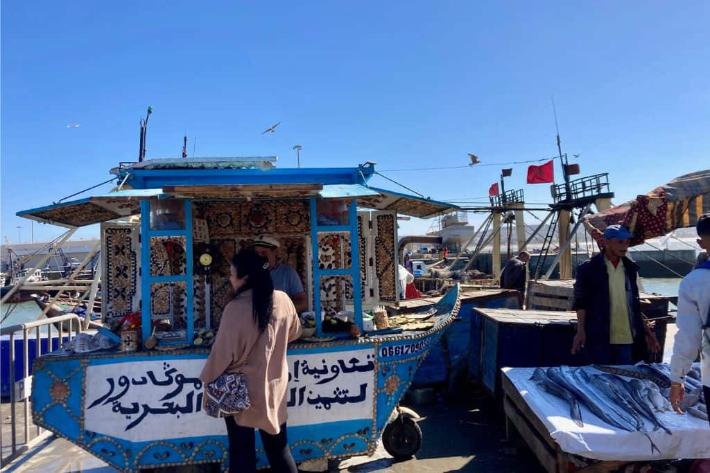 A woman is stood looking at a fish stall with her back to the camera. She is a at fish market and there are other stalls either side of her. This image is taken in Essaouira which is well worth visiting.
