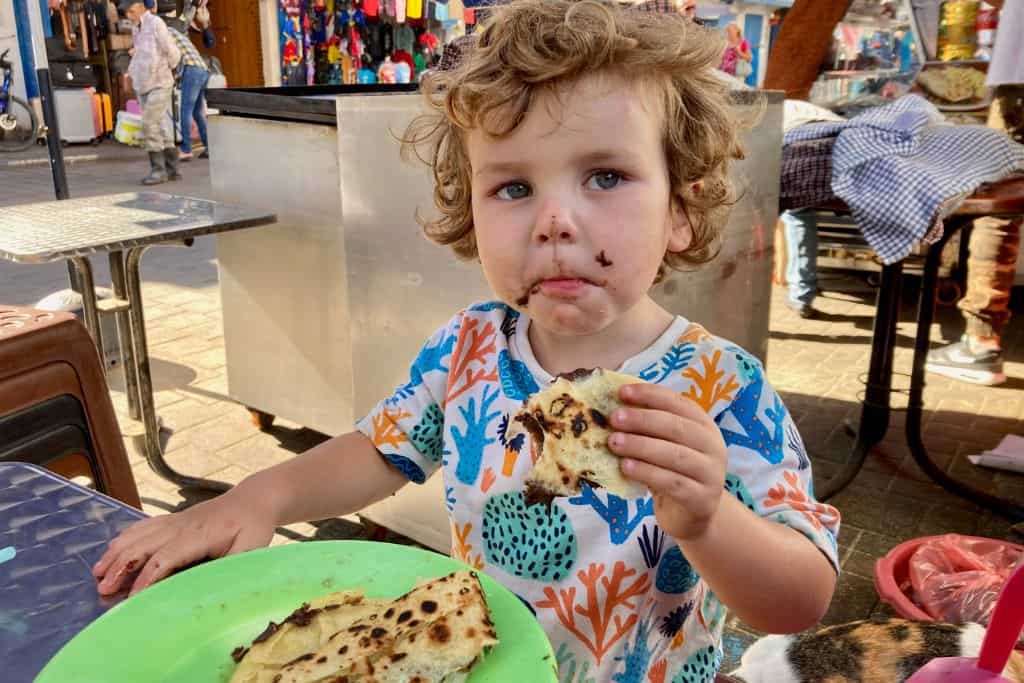 A little boy in a brightly coloured is looking at the camera with a cheeky look on his face as he has chocolate around his mouth from eating a crepe.  