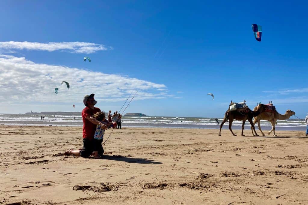 The photo is taken on a beach in Essaouira which is well worth visiting. It's very windy and in the distance are some kite surfers. In the middle of the image is the side on profile of a man on his knees looking at the sky as he flies a kite. Stood on his lap is a 3 year old boy who is helping to fly the kite.