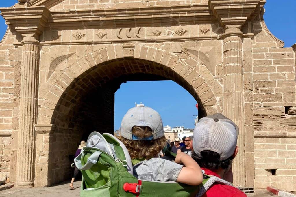 There is a large fort gate in the centre of the image which leads from the fishing port in Essaouira.  In the front of the gate is a man with his back to the camera looking at the gate, and on his back in a carrier is his son who is also looking at the gate.