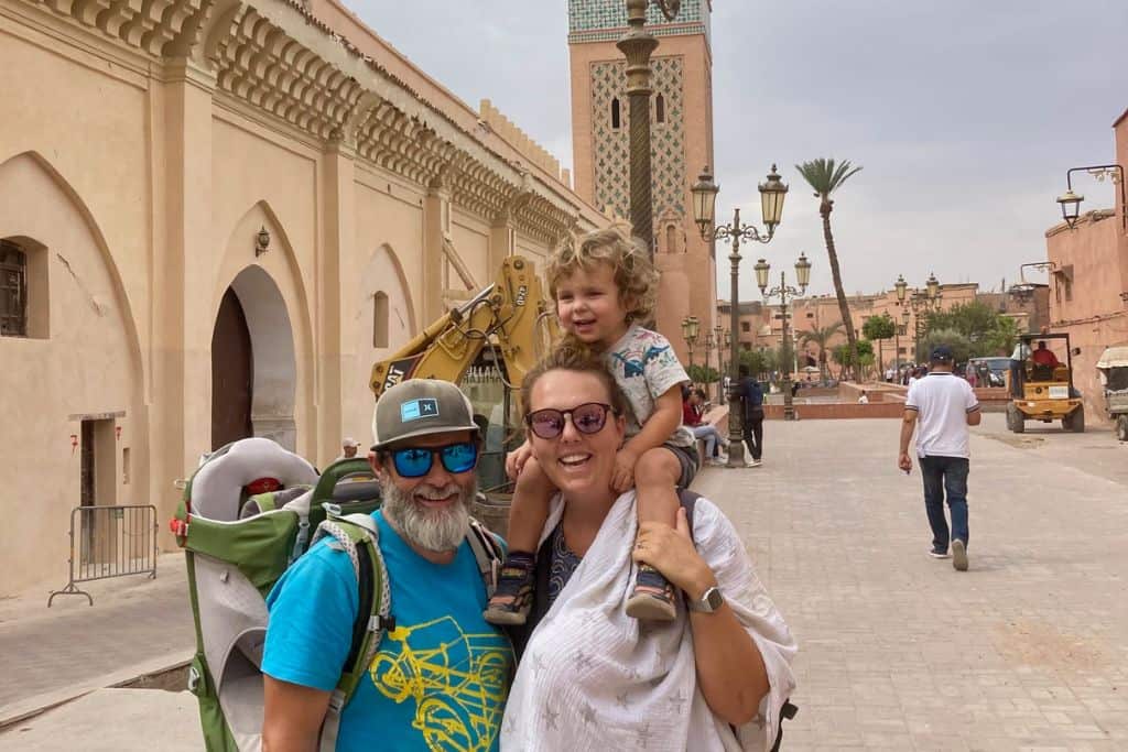 A man in a turquiose t-shirt, sunglasses and baseball cap is looking at the camera and smiling.  On his back is a child carrier backpack which is empty. Next to the man is a woman who has their son on her shoulders, and on her front covered by a muslin is her baby in a baby carrier. Behind them is Marrakech in Morocco which is safe for families.