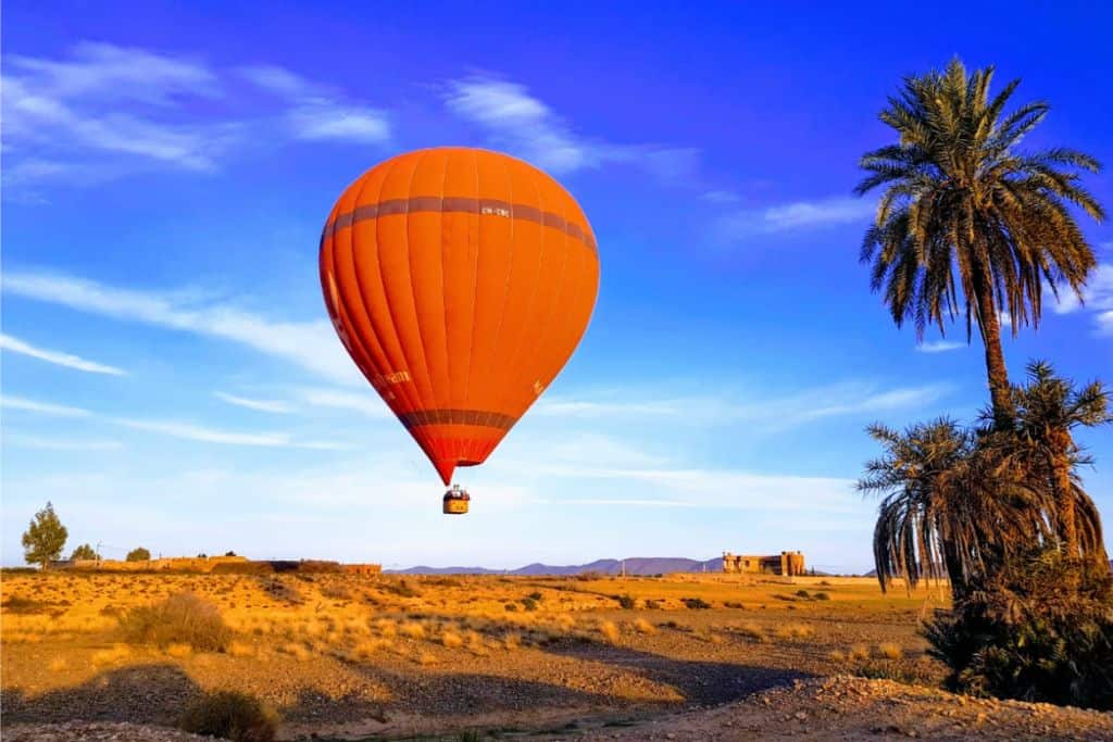 A bright blue sky with an orange hot air balloon in it. To the right are some palm trees and in the lower background is the desert in Marrakech.
