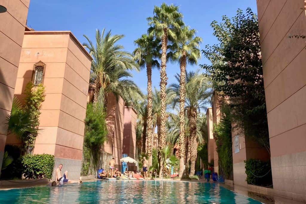 A hotel swimming pool in Marrakech in Morocco with kids playing in it. To the sides are the buildings of the hotel and in the middle are some very large tall palm trees.