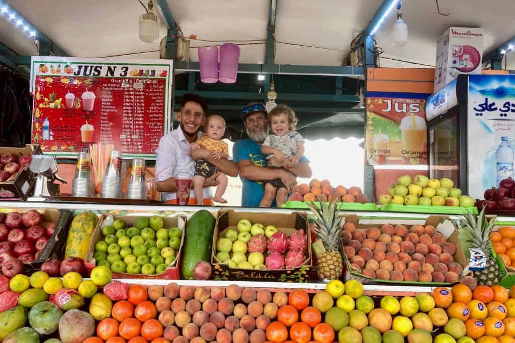 A man in a blue t-shirt is holding his three year old son, and next to him is another man in a white shirt that is holding a baby. They are in a juice stand in Marrakech with kids. In front of them is boxes of different fruits ready for them to juice.