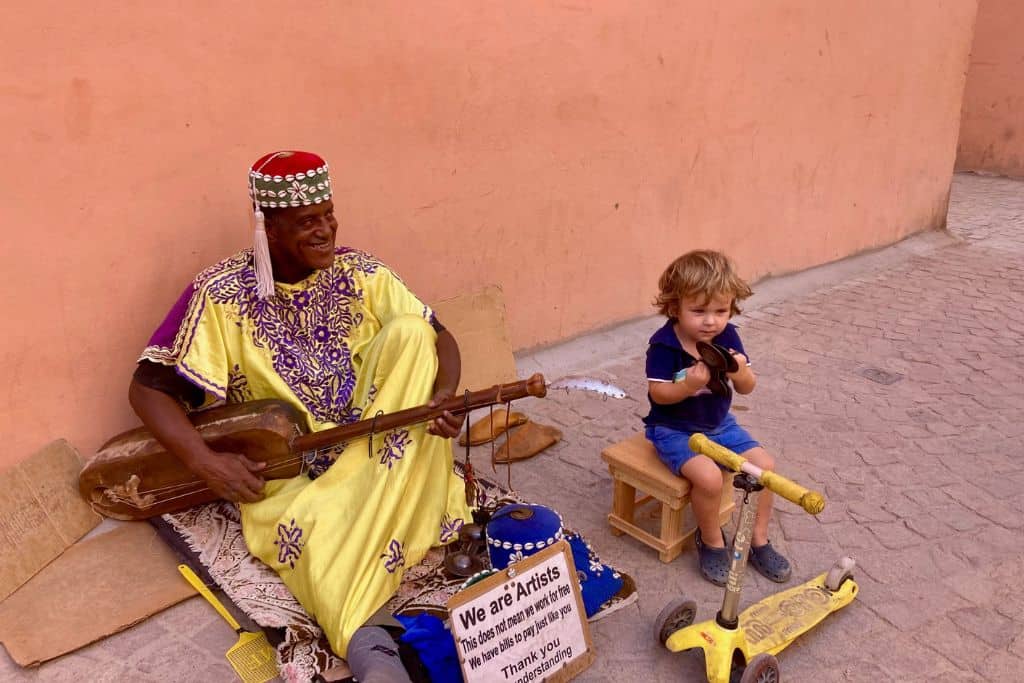 A Moroccan man is playing a guitar sat on the floor and has on a traditional outfit in yellow and a fes on his head. Next to him is a little boy that is holding some finger chimes and is playing along with him. They are in Marrakech in Morocco and the man is a street performer playing local music.  The boy is happily sitting there as Morocco is very safe for families.
