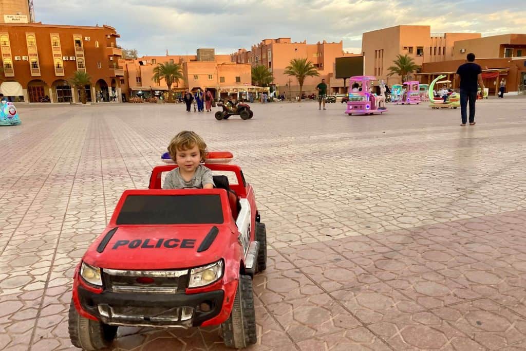 A little boy is facing the camera sat in a red electric police car that he's driving in a square in Tinghir in Morocco.