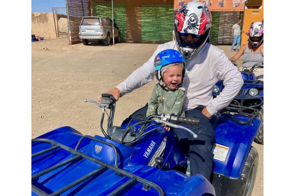 A man in a long sleeved white t-shirt has on a motorcycle helmet and is sat on a blue quad bike. In front of him in a green long sleeved t-shirt is his three year old son. They are on the edge of the Erg Chebbi desert as they are about to quad bike in the sand dunes.  This is a safe activity for families to try in Morocco.