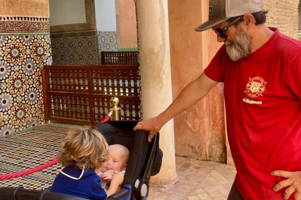 A man is a red t-shirt has his hand on a pram which has his baby son in it. The baby is facing the camera and in front of his looking at him with his back to us is his brother who is playing with them. In the background are the Sadiaan Tombs in Marrakech that you can visit with kids.