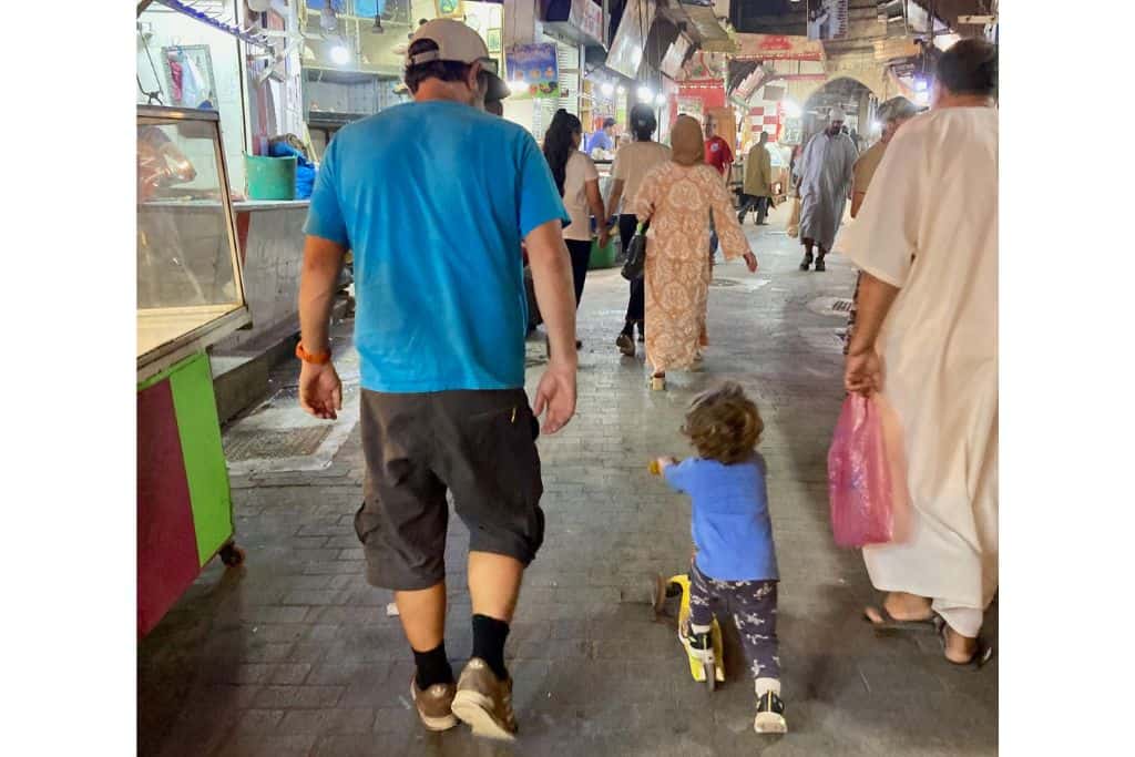 There is a man with a blue t-shirt and his back to the camera. Next to him is a little boy in a blue t-shirt who is riding a scooter in the souk in Marrakech.