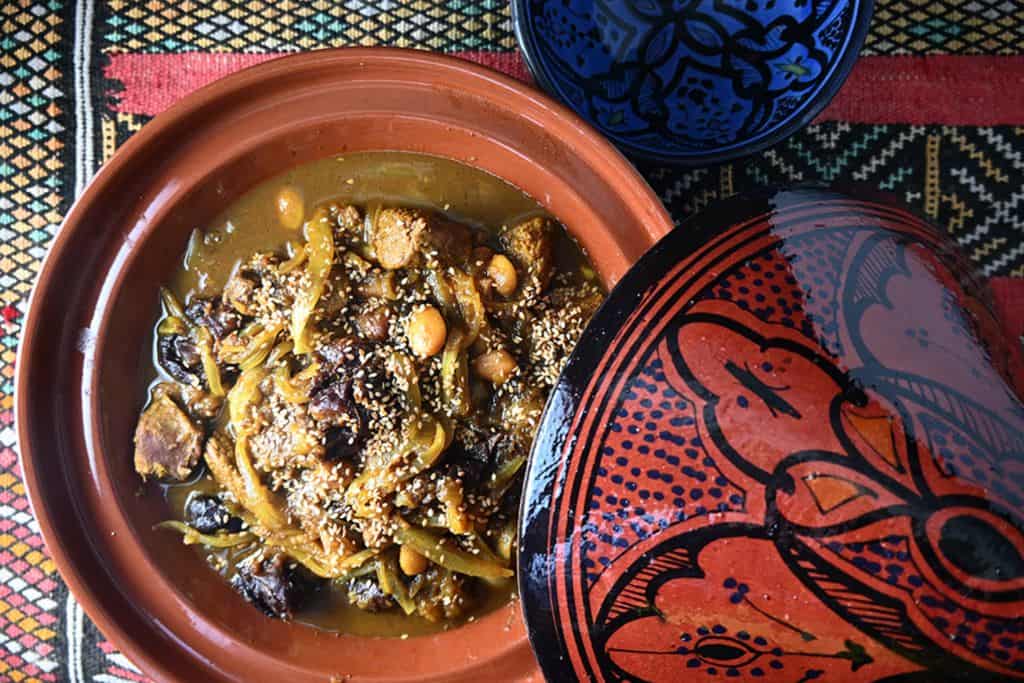 A clay dish is holding a tagine in it.  To the side of the base of the dish is the lid which is a dark orange colour with a black design painted on it. 