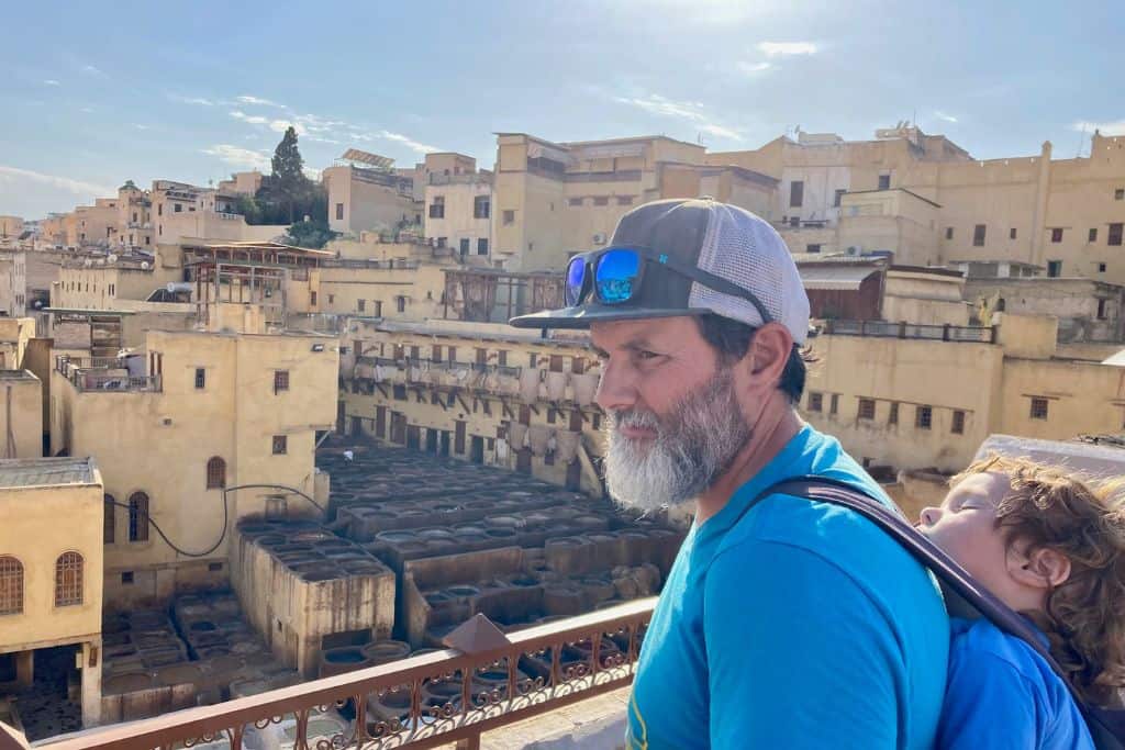 Close-up of a man with a blue t-shirt on and a baseball cap with sunglasses on it is looking to the left.  On his back in a carrier is his toddler son who is asleep.  In the background are the tanneries in Fes which is where he is travelling with his kids.
