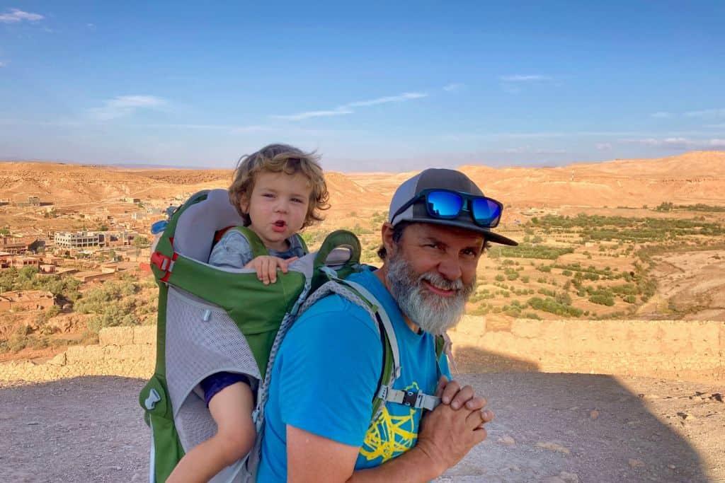 Man in a blue t-shirt and a baseball cap with sunglasses on his head is wearing a child carrier on his back with his son in it. Behind them is Ait Benhaddou in Morocco.