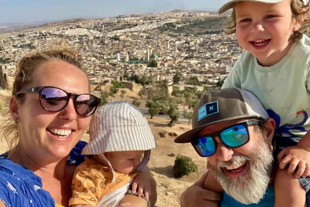 Close up of a mum with blue shirt and sunglasses on holding her baby with a sun hat on. Next to here is her boyfriend who has sunglasses and a hat on and has their son on his shoulders.  In the background is the city of Fes in Morocco that they are travelling through with their kids.