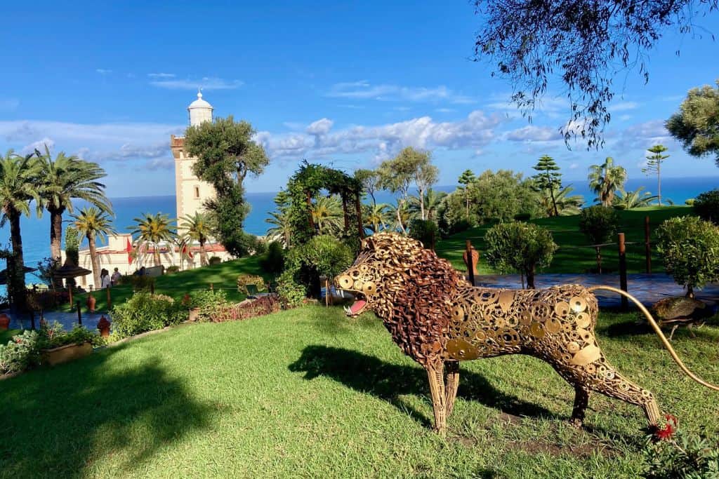 A photo of a garden with a metal statue of a lion roaring on the grass.  Behind the lion are some bushes and then behind them is the lighthouse at Cap Spartel.
