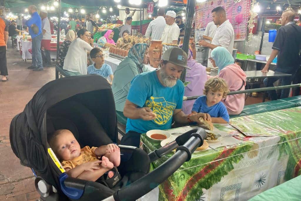 A man in a blue t-shirt is sat at a table in a food stall with his son also in a blue t-shirt next to him.  On the otherside in a pram is his baby in an orange t-shirt. They are eating food in the main square of Marrakech.