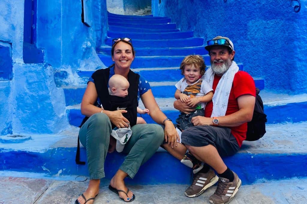 A family made up of a mum and dad with their kids are sat on the blue steps in the city of Chechaouen in Morocco.  The mum has a baby in a baby carrier o her, and the dad in a red t-shirt, baseball cap has his 3 year old son on his lap.  the steps and walls are an azure blue.