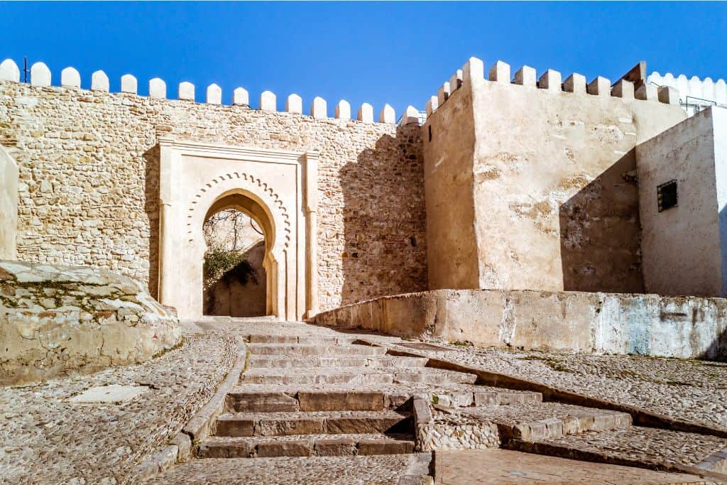 The Medina gate in Tangier which is worth visiting. The sand coloured stone wall with gate in it has some stairs leading up to it.  The sky is blue behind it.
