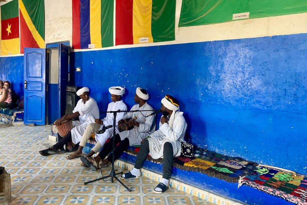 Four African men in long sleeved long shirts with white turbans are performing music as part of a Gaou band. They are sat on a seat in front of a blue wall and above the wall are several flags from the Sahara.