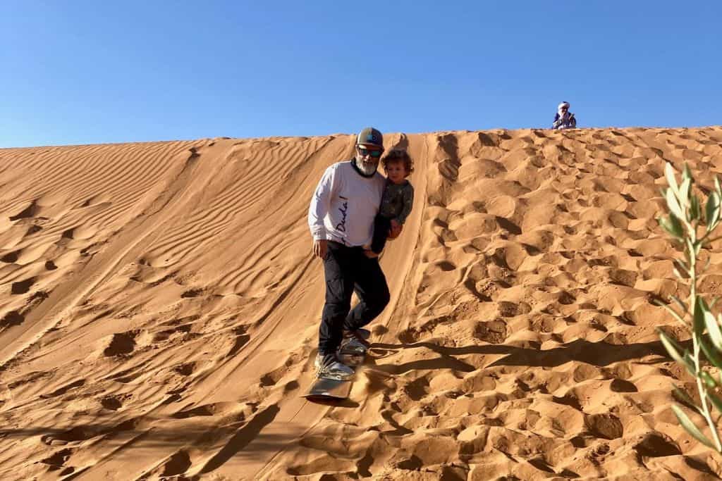 A man has his three year old son in his arms and is half way down a sand dune on a sand board wit his son.