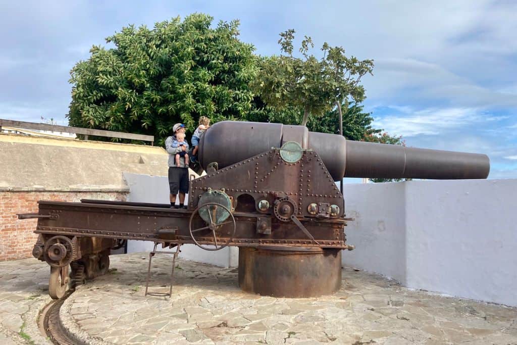 A very large mechanical cannon is being pointed over the city walls in Tangier. There is a man holding his 3 year old son so that he can pretend to be firing the cannon. 