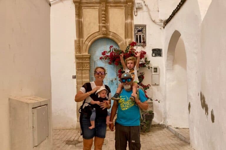 A family is at the Kasbah in Rabat, the mum is wearing a white top and on her front is her baby in a carrier. The man has a blue t-shirt on and his son on his shoulders. They are all smiling at the camera and behind them is a blue wooden door within a stone arch. Visiting the kasbah is one of the best things to do in Rabat.